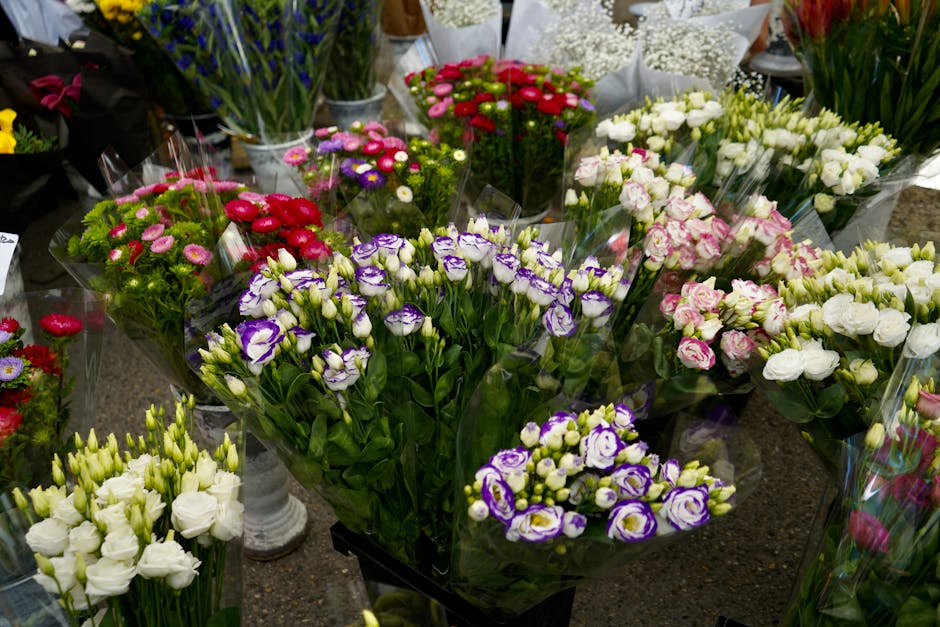 A variety of fresh flower bouquets displayed on a market table, featuring purple and white lisianthus, white eustoma, and pink and white roses, all wrapped in transparent plastic. The flowers are arra