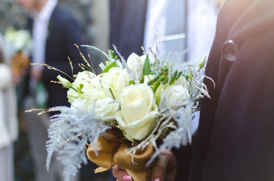 A person dressed in a dark suit is holding a bouquet consisting of white roses and other white flowers, accented with delicate silver-gray foliage and wispy decorative elements. The bouquet is wrapped