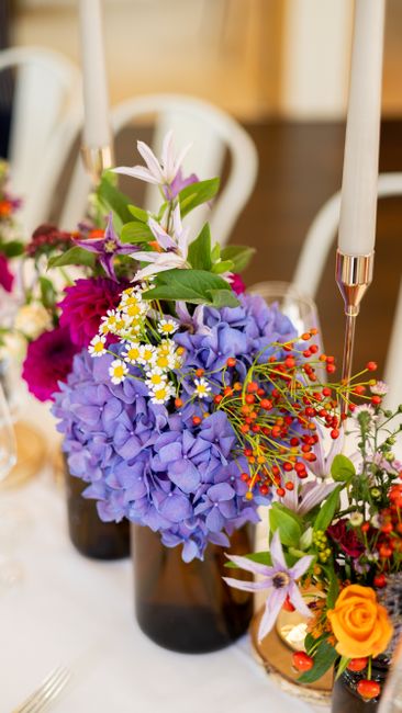 A vibrant floral arrangement featuring a cluster of purple hydrangeas, complemented by small white daisies with yellow centers, pink and purple clematis with elongated petals, bright orange roses, and