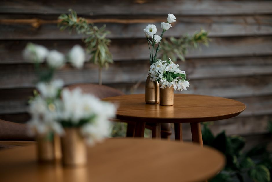 A floral arrangement styled for the Roundhouse event, featuring white lisianthus, delphiniums, and hydrangeas in a small, metallic bronze vase. The bouquet is placed on a polished wooden table, with n