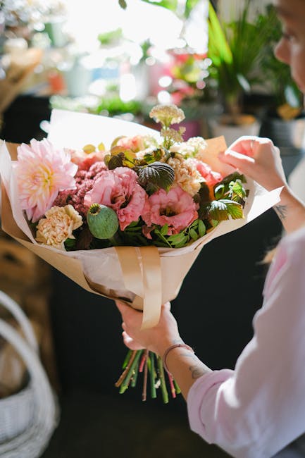 A person holding a freshly prepared floral bouquet featuring large pink peonies, pale yellow carnations, and white filler flowers wrapped in white tissue paper and tied with a beige ribbon. The arrang