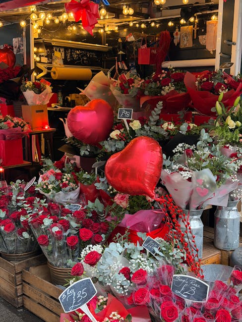 A vibrant floral display at a flower shop features an assortment of fresh red roses, white baby's breath, and green foliage arranged in baskets and wrapped in festive pink and red paper. Two large, sh