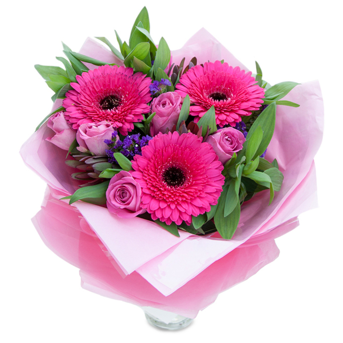 The composition opens with three striking fuchsia Gerbera daisies placed like focal points, their saturated petals forming perfect concentric rings around dark, almost bronze centers that punctuate the palette with graphic contrast. Nestling between these larger blooms are roses in a muted lavender-pink or dusty-rose shade, their petals tightly furled in youthful spirals that suggest freshness and intimacy. Between the major forms, small clusters of purple statice float on filigree stems, introducing a cooler violet accent and a papery texture that contrasts with the smooth rose petals. A generous spray of greenery-broad salal-like leaves and slim eucalyptus stems-frames the flowers, the greens slightly glossy and emphasizing the vibrancy of the pinks. The wrapping is deliberately elegant: a soft, matte pale pink tissue wraps the stems, while an outer layer of translucent fuchsia-pink cellophane, with a faint crinkle, lends a delicate gleam and completes the monochromatic theme. The lower edge reveals the rim of a clear glass vase, implying the bouquet is arranged for immediate placement on a side table, mantel, or bedside. In terms of scent and atmosphere, imagine a gentle rose perfume softened by green notes from the foliage and the clean, airy quality of statice; it conjures a quiet morning near Chalk Farm Road or a cheerful afternoon after browsing Camden Market. Intended for celebrations, tokens of affection, or to lift someone's spirits, this bouquet balances structured floristry with spontaneous colour, speaking of thoughtful craftsmanship and local care.